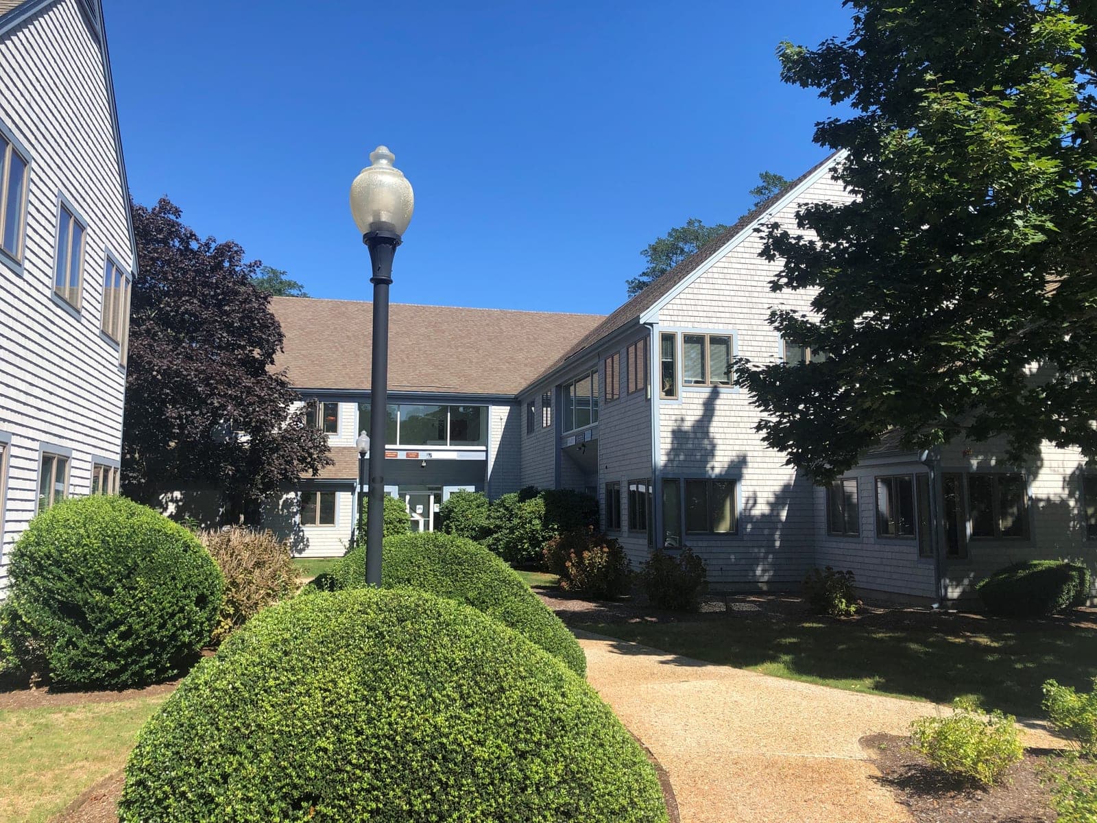 The office building at 19 Bay State Ct in Brewster, where Cape Gynecology is located — a two-story grey-shingled Cape-style building with blue-grey window trim, set behind rounded boxwood hedges along a paved walkway.
